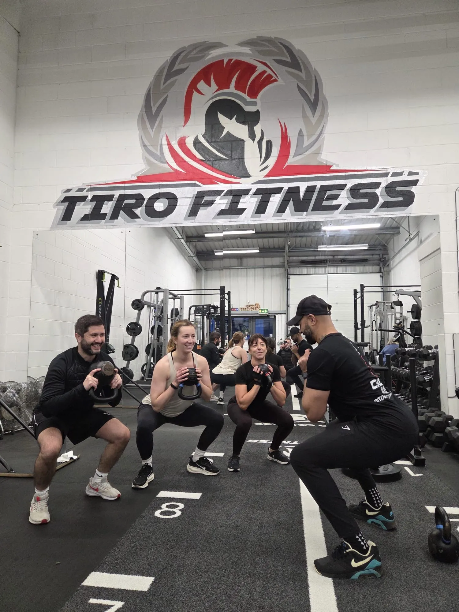 Group of members doing kettlebell goblet squats at Tiro Fitness, smiling under the gym's wall logo