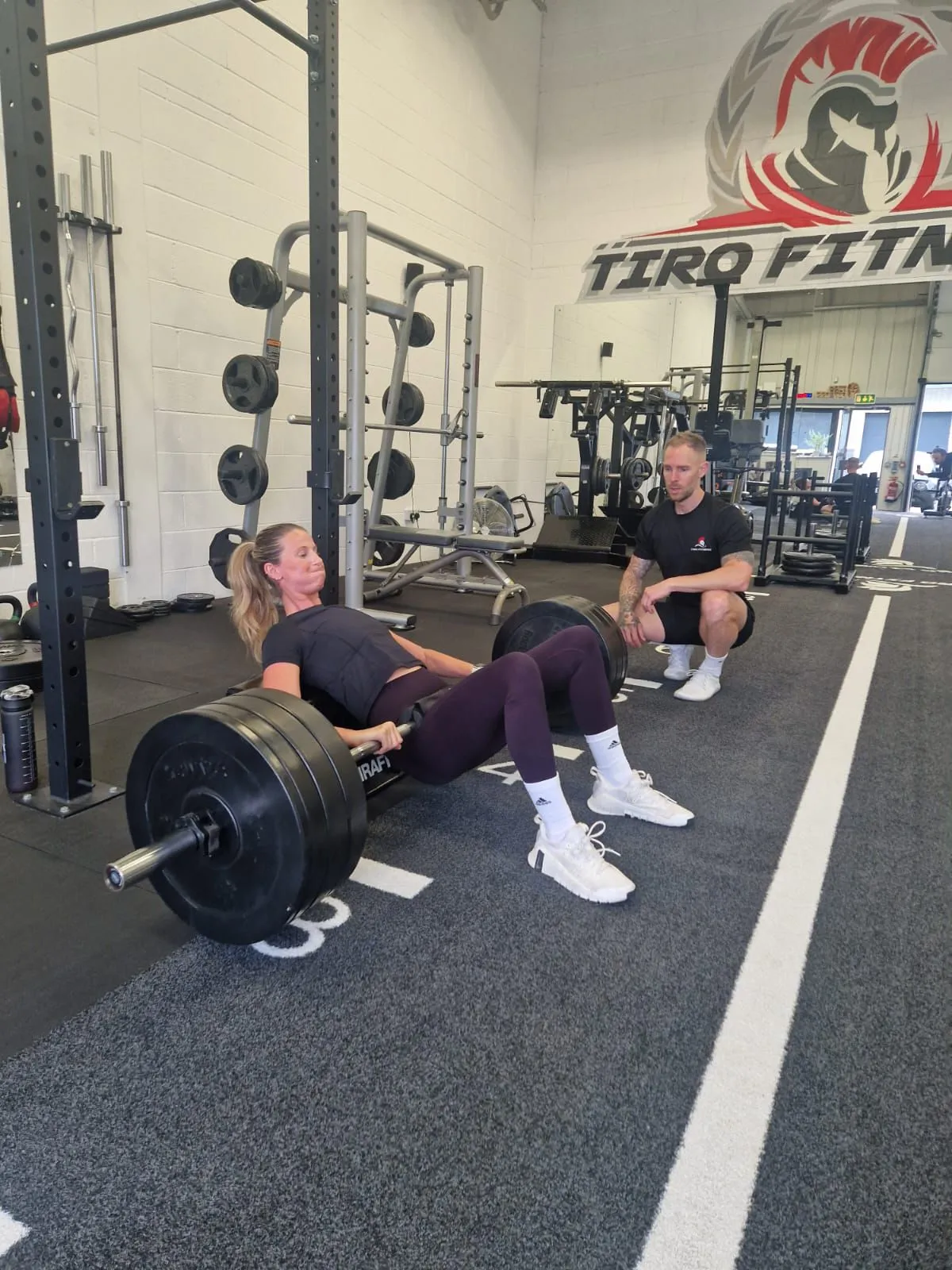 Woman performing barbell hip thrusts with a trainer coaching at Tiro Fitness