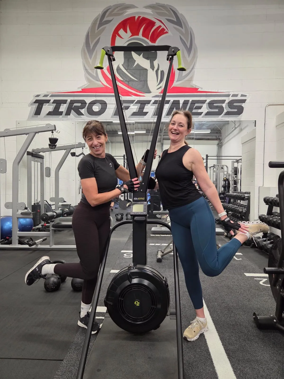 Two women laughing together while posing with a ski erg at Tiro Fitness